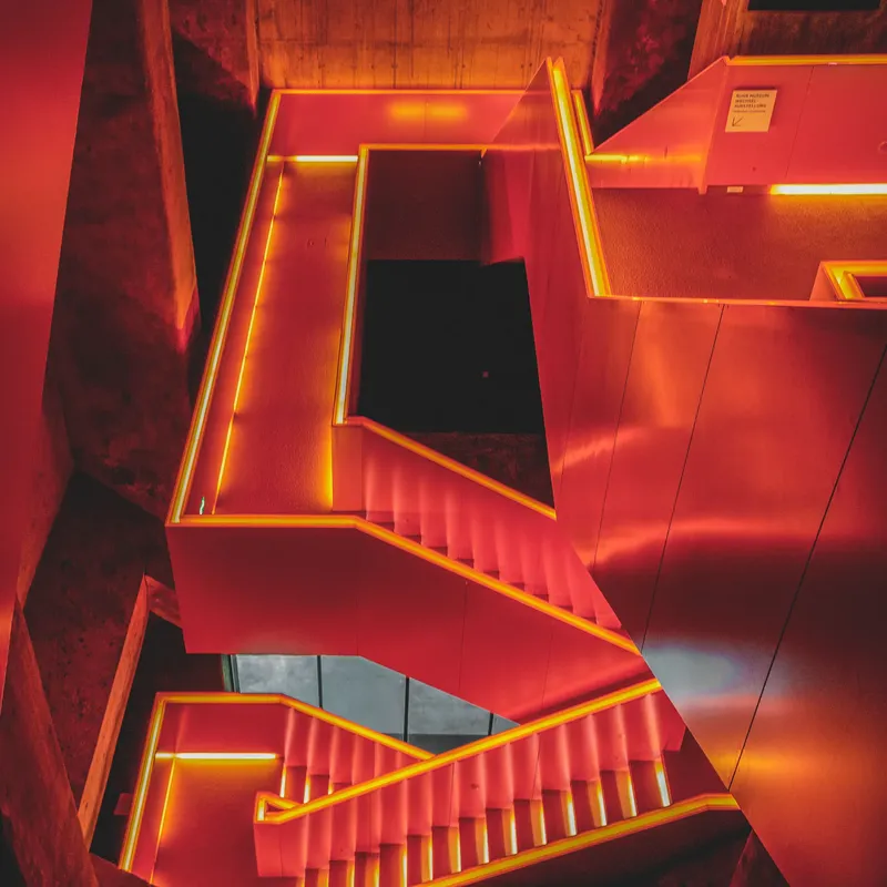 Moderne Architekturaufnahme einer rot beleuchteten Treppenanlage mit geometrischem Design und LED-Akzentbeleuchtung in Orange. Minimalistisches Interior Design mit dramatischer Farbgebung und perspektivischer Komposition.
