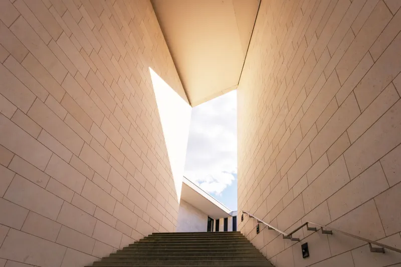 Elegantes modernes Treppenhaus mit beigen Steinwänden und dramatischem Lichteinfall. Minimalistische Architektur-Fotografie zeigt klare Linien, geometrische Formen und ein harmonisches Zusammenspiel von Licht und Schatten in urbaner Gebäudearchitektur.
