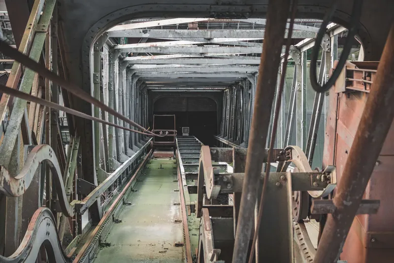 Atmosphärische Industriefotografie eines historischen Bergbautunnels mit original erhaltener Fördertechnik. Der symmetrisch komponierte Gang zeigt eindrucksvoll die industrielle Architektur mit Stahlträgern, Förderband und mechanischen Elementen in authentischer Patina.
