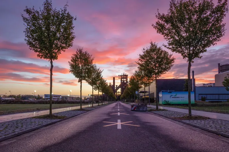 Atemberaubender Sonnenaufgang über der Industriekulisse Phoenix West in Dortmund. Symmetrisch angelegte Baumallee führt zum historischen Hochofen, umrahmt von einem spektakulären Himmel in Rosa- und Orangetönen. Perfekte Symbiose aus Industriekultur und Naturschauspiel.
