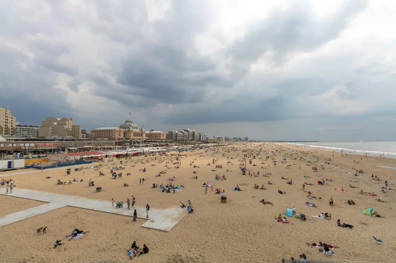 Panoramablick über den Strand von Scheveningen, Den Haag: Imposantes historisches Kurhaus-Hotel, weitläufiger Sandstrand mit Besuchern, moderne Strandpromenade und dramatische Wolkenformation über der Nordseeküste. Authentische Aufnahme des beliebtesten niederländischen Seebads.
