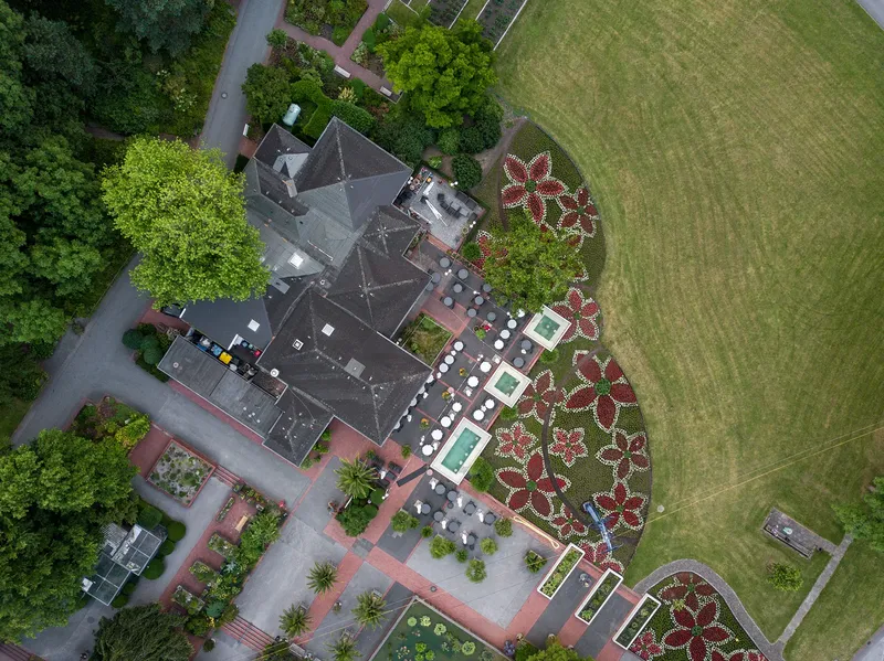 Luftaufnahme einer exklusiven Gartenanlage mit kunstvoll gestalteten Blumenbeeten in Form von roten Blüten, drei dekorativen Wasserbecken und einer großzügigen Terrasse mit Sitzgelegenheiten. Die Drohnenaufnahme zeigt die perfekte Symbiose zwischen architektonischer Gestaltung und Landschaftsgärtnerei von oben.
