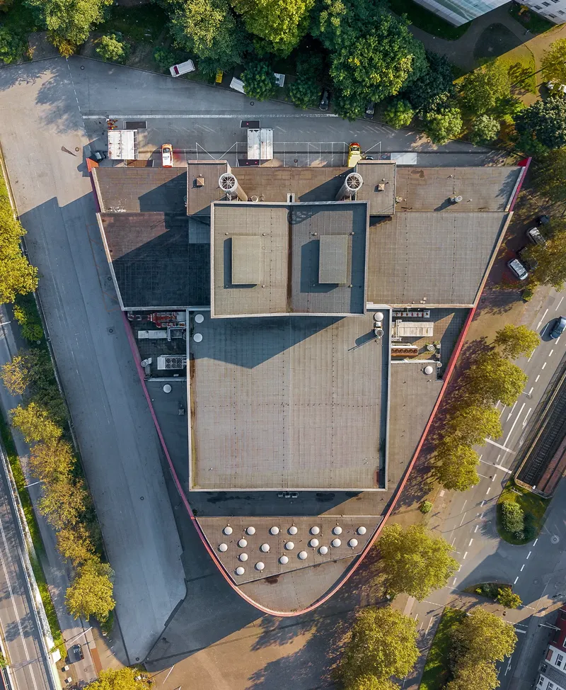 Luftaufnahme vom Marientheater Duisburg mit charakteristischer Spitzform, technischen Dachaufbauten und kreisförmig angeordneten Installationen, eingebettet in urbane Infrastruktur mit Parkflächen und Grünanlagen. Drohnenfotografie zeigt architektonische Details aus der Vogelperspektive.
