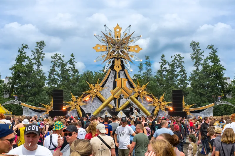 Defqon1 Gold Stage mit imposanter goldener Stern-Architektur und LED-Beleuchtung. Atmosphärische Aufnahme eines Open-Air Musikfestivals mit begeistertem Publikum vor naturnaher Kulisse. Festival ElectronicMusic EventPhotography
