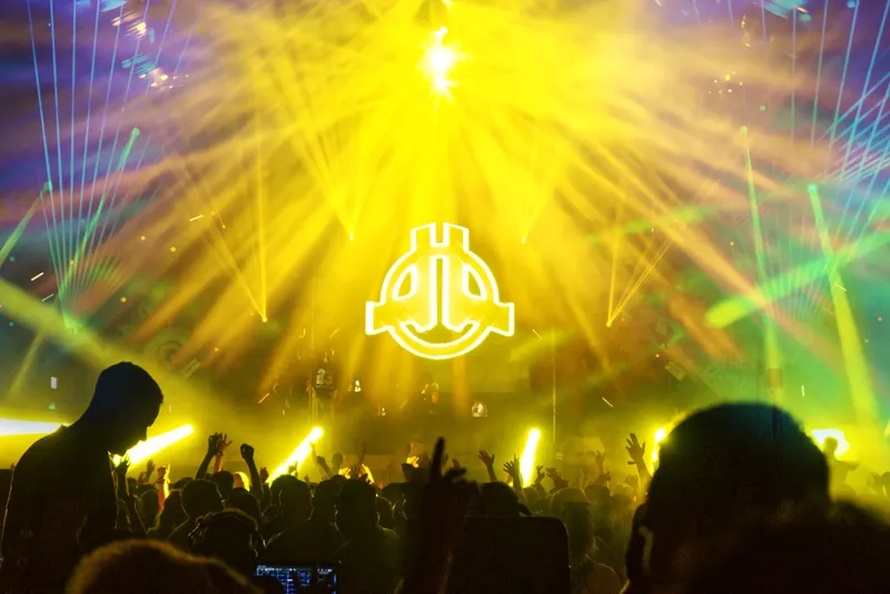 Spektakuläre Konzertaufnahme eines EDM-Events mit beeindruckender Lichtshow in Gelb- und Grüntönen. Energiegeladene Atmosphäre mit jubelndem Publikum vor einer großen Bühne mit charakteristischem DJ-Logo und modernster Laser-Technik. Authentische Festival-Stimmung eingefangen in 1536x1026 Pixeln.
