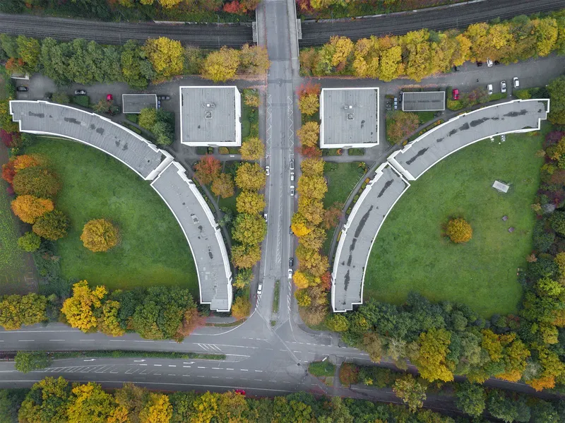 Beeindruckende Luftaufnahme einer symmetrischen Architektur mit zwei geschwungenen Gebäudekomplexen, eingebettet in herbstliche Landschaft mit goldgelben Bäumen, Bahngleisen und Straßeninfrastruktur. Ein perfektes Beispiel moderner Stadtplanung und nachhaltiger Integration in die Natur.
