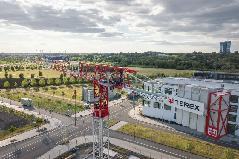 Luftaufnahme des Terex Abrams Krane Firmengeländes mit imposantem roten Turmdrehkran im Vordergrund. Moderne Industriearchitektur mit weißem Firmengebäude, weitläufiger Infrastruktur und Grünflächen. Im Hintergrund ist das Stadtbild mit Sportstadion und Hochhaus zu sehen.

