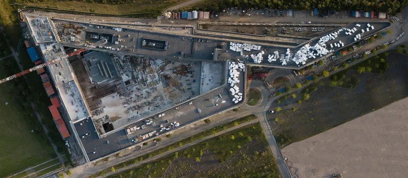 Luftaufnahme einer industriellen Großbaustelle mit modernen Gebäudekomplexen, Lagerflächen und logistischer Infrastruktur. Die Drohnenaufnahme zeigt die weitläufige Anlage mit weißen Containern, Baugerüsten und angrenzenden Grünflächen aus der Vogelperspektive.
